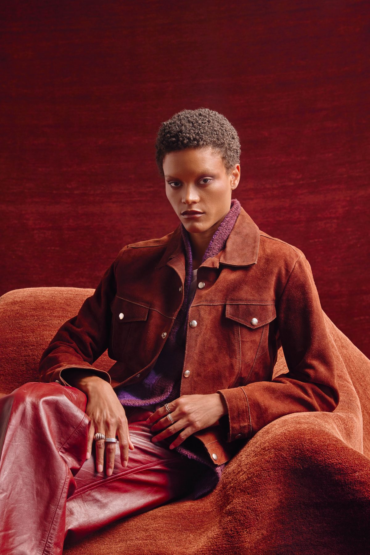 A model in a brown suede jacket, purple knit vest and red leather pants sits in an orange upholstered chair before a red Armadillo rug.