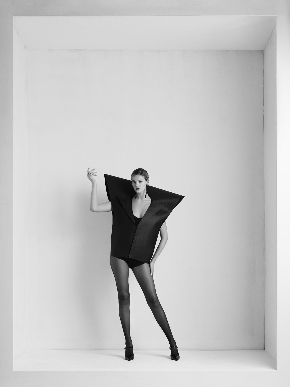 A model in a black Ome Epōt Fold jacket with wide angular points and sheer tights poses in a white alcove, in black and white.