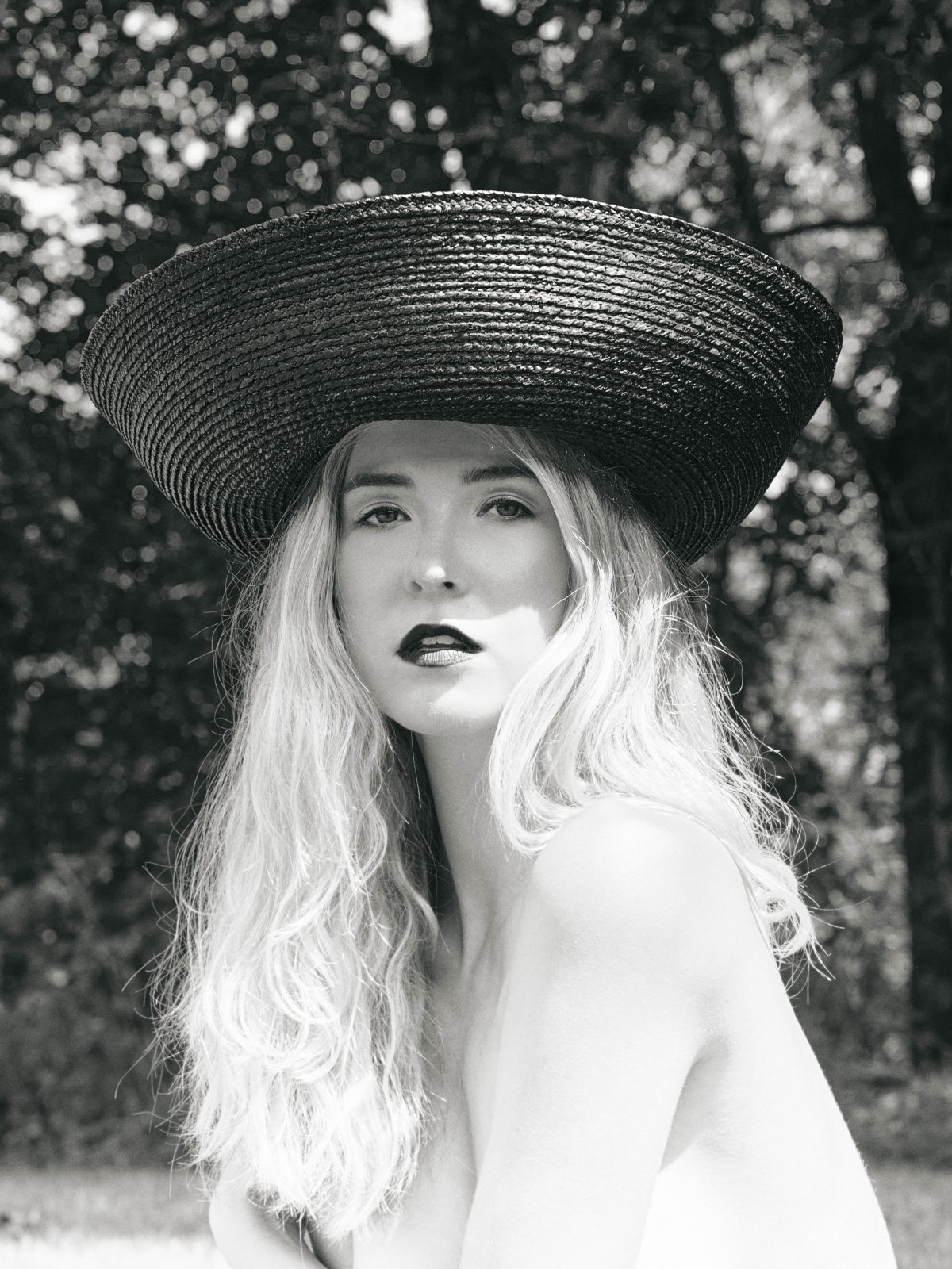 A model in a wide woven Helen Kaminski hat, with long blond hair and dark lipstick, bare-shouldered in front of trees, in black and white.