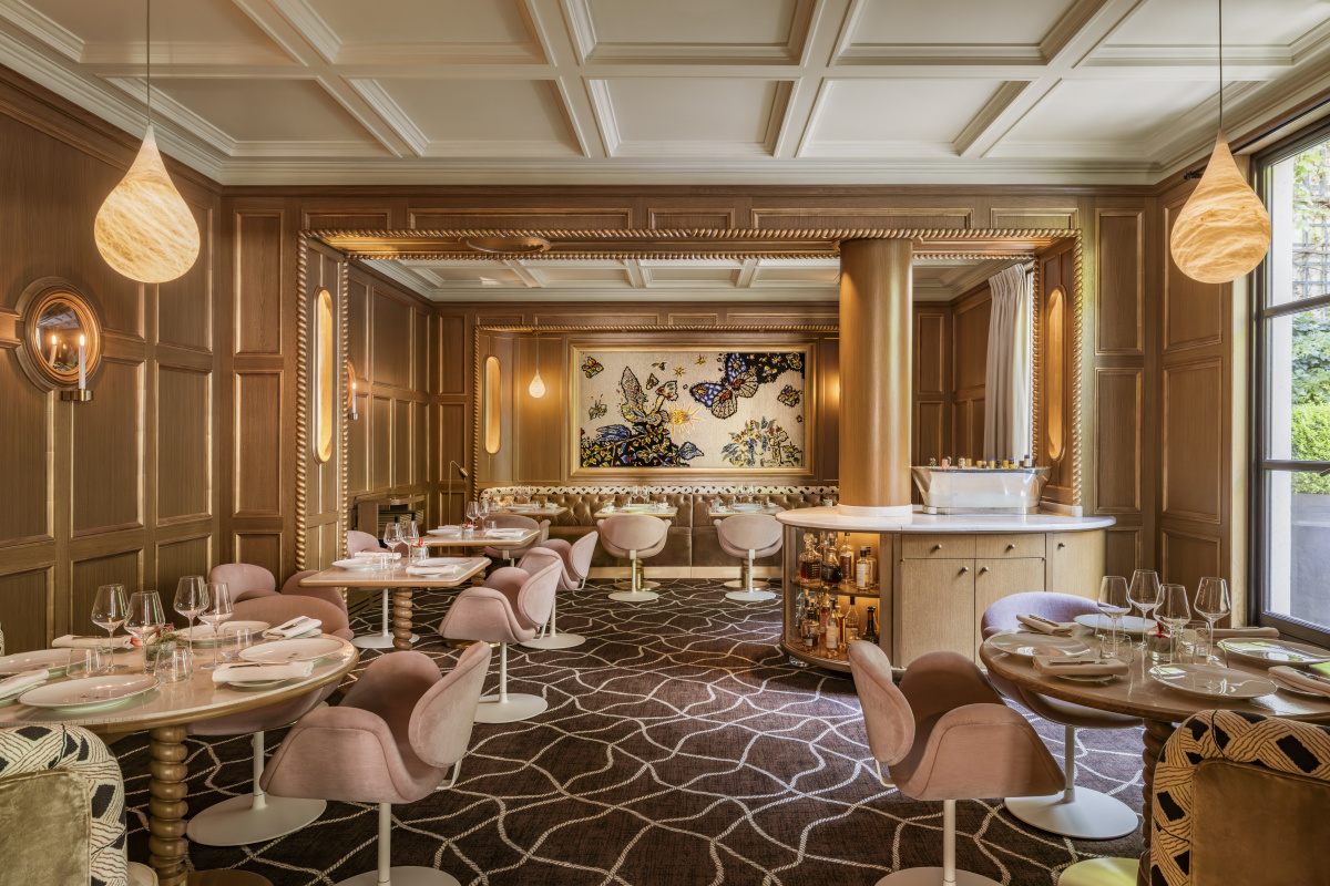 The dining room of restaurant Anne at the Pavillon de la Reine, with oak paneling, gilded moldings, pink velvet chairs, a butterfly tapestry, and a patterned brown carpet.
