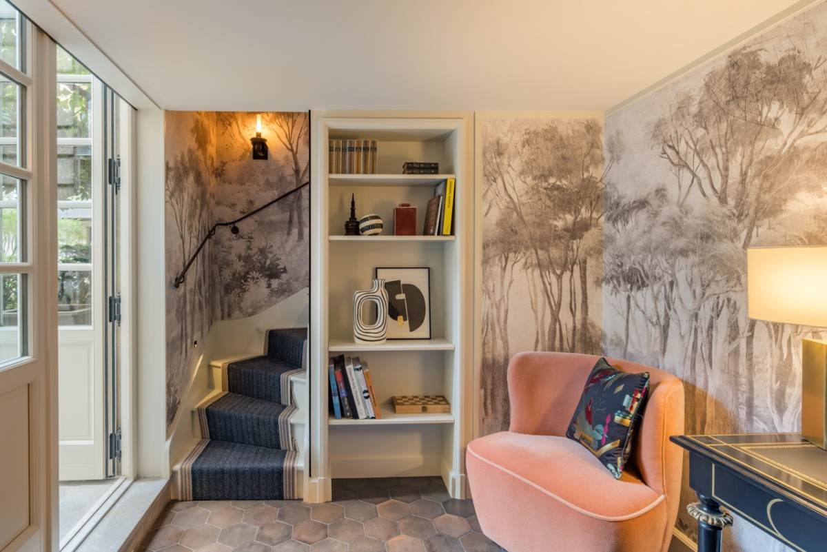 A stair hall at the Pavillon de la Reine with a sepia tree mural, a navy stair runner, built-in bookshelves, hexagonal terracotta floor tiles, and a peach velvet armchair.