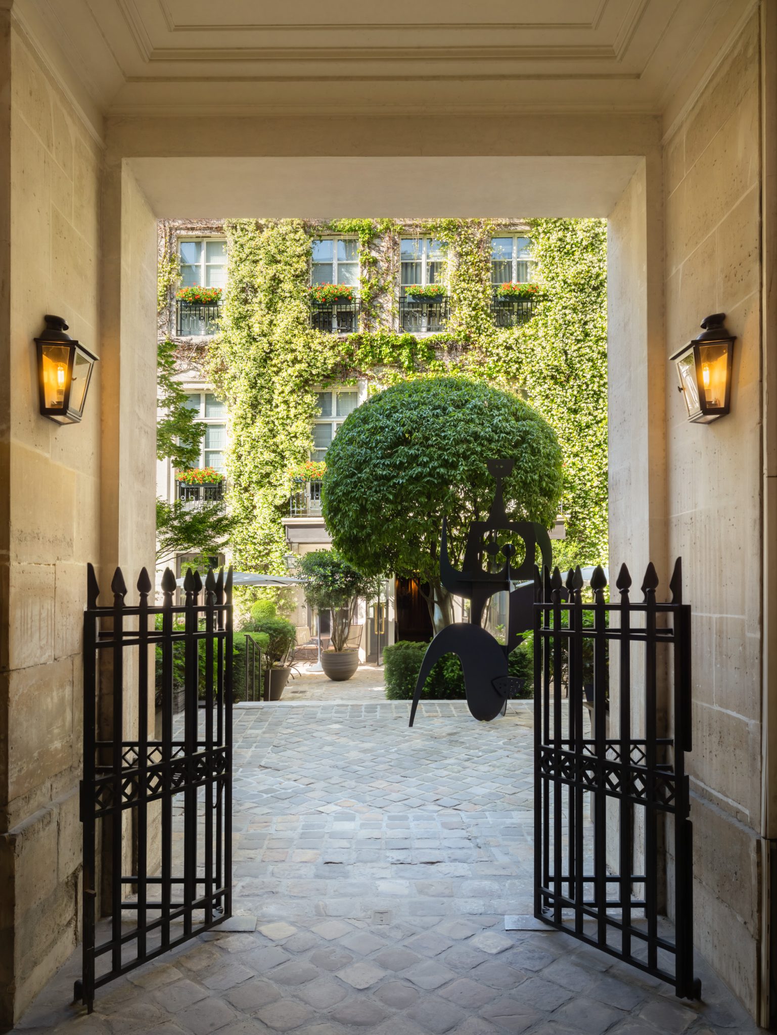 Open iron gates in a stone passageway leading to a cobbled courtyard with a sculpted tree and the ivy-covered facade of the Pavillon de la Reine in P