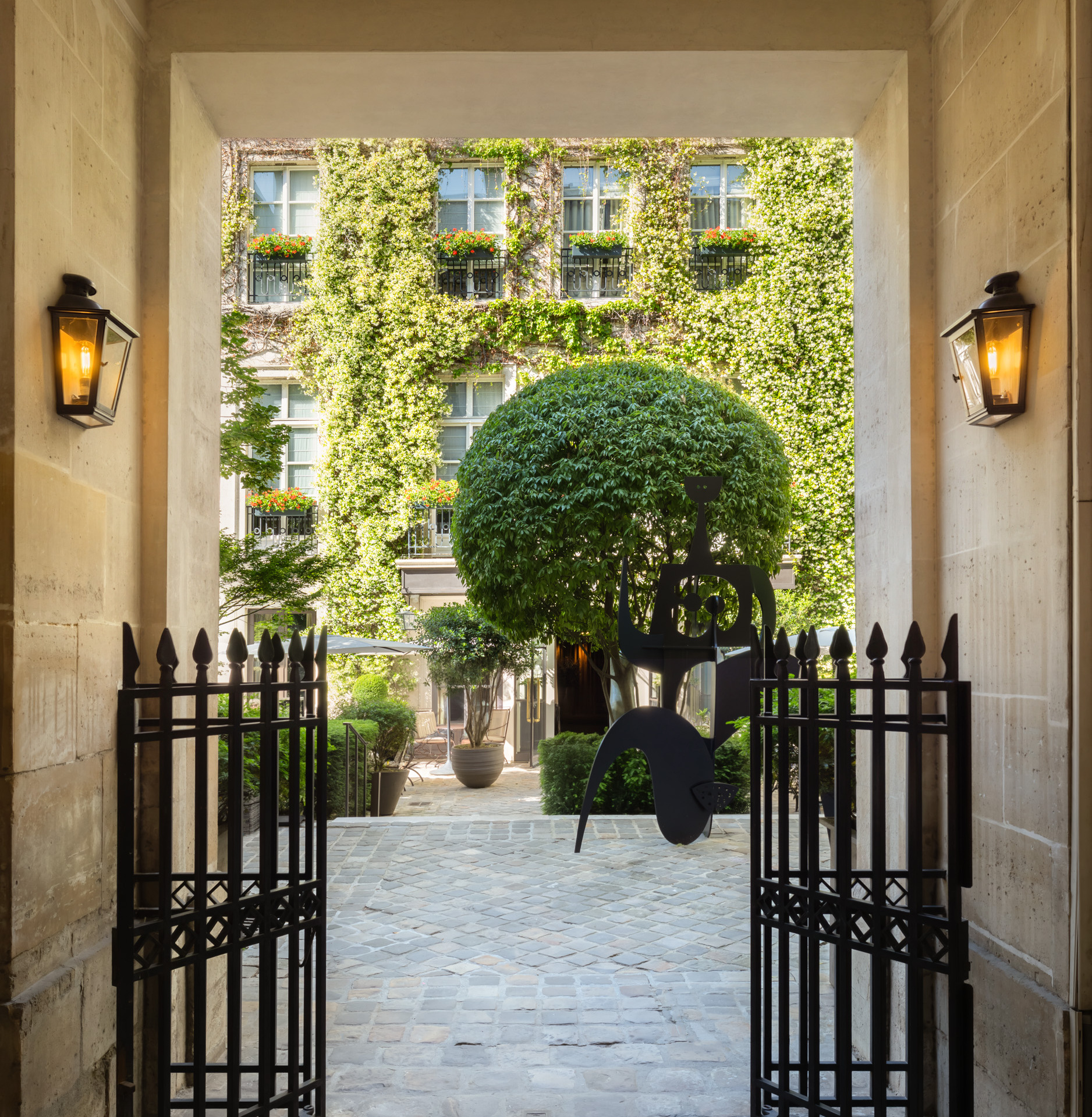 Open iron gates in a stone passageway leading to a cobbled courtyard with a sculpted tree and the ivy-covered facade of the Pavillon de la Reine in P