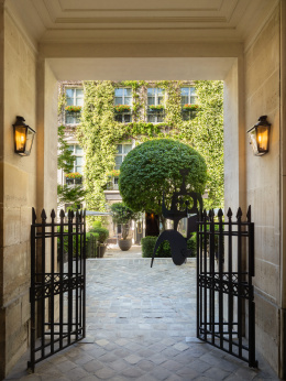 Open iron gates in a stone passageway leading to a cobbled courtyard with a sculpted tree and the ivy-covered facade of the Pavillon de la Reine in P