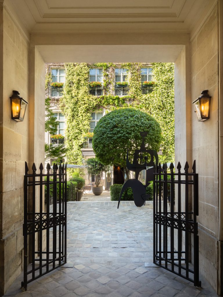 Open iron gates in a stone passageway leading to a cobbled courtyard with a sculpted tree and the ivy-covered facade of the Pavillon de la Reine in P