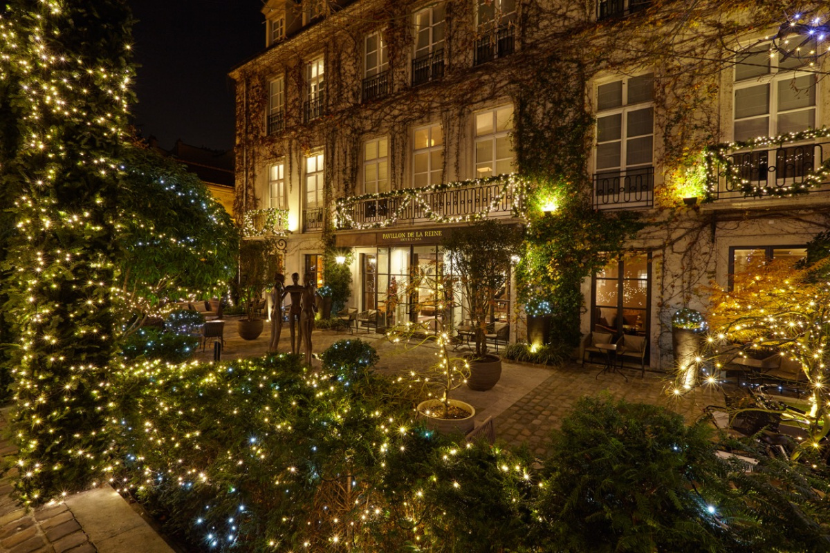The Pavillon de la Reine's courtyard at night in winter, with string lights in the garden, garlands on the balconies, and a bronze sculpture of three figures before the ivy-covered facade.