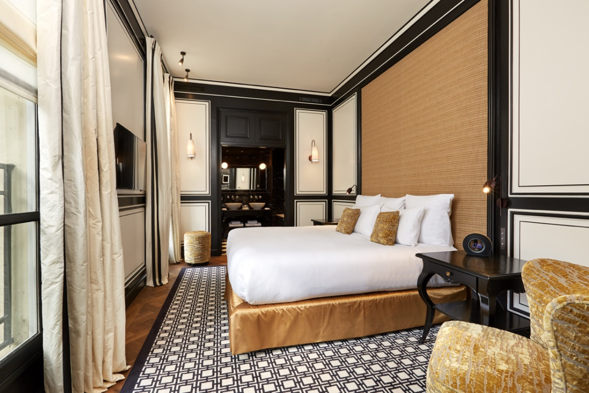 A contemporary guest room at the Pavillon de la Reine with black-and-white paneled walls, a raffia headboard, a gold silk bed skirt, a geometric rug, and a dark marble bathroom through the doorway.