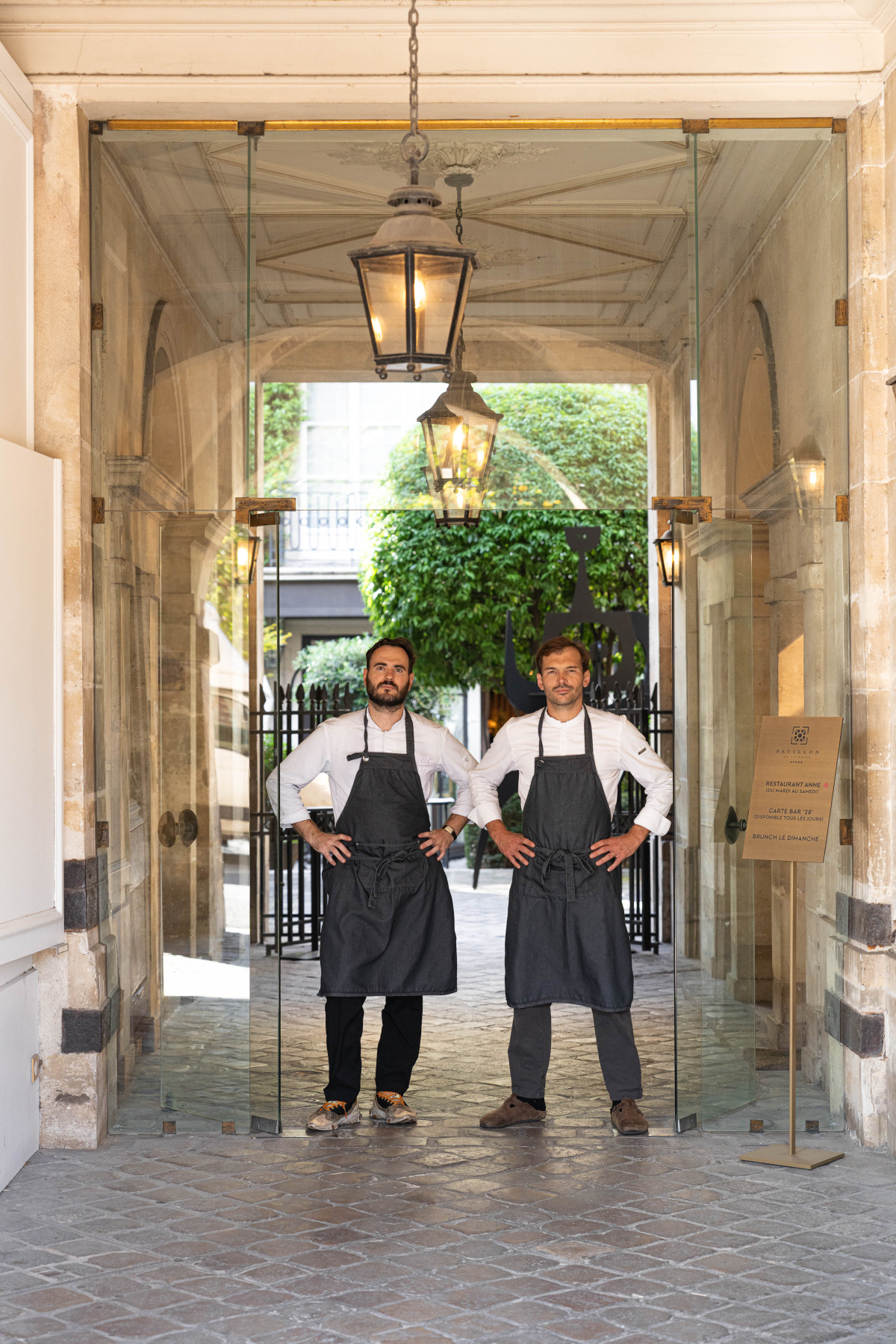 Two chefs in gray aprons standing in a stone entry passage with lanterns and glass doors, the courtyard of the Pavillon de la Reine visible behind them.
