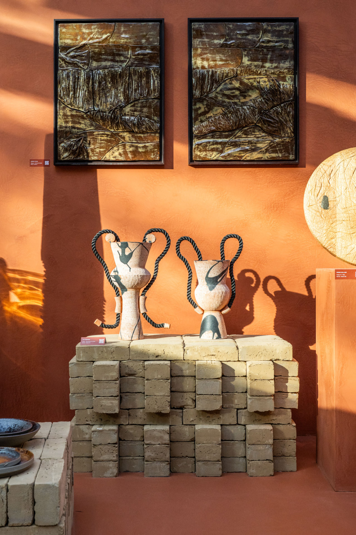 Two stoneware urns with rope handles on a brick plinth against a terracotta wall