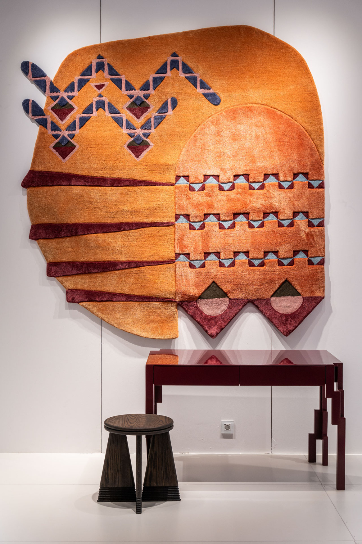 Orange tufted wall hanging with geometric pattern above a dark red desk and stool