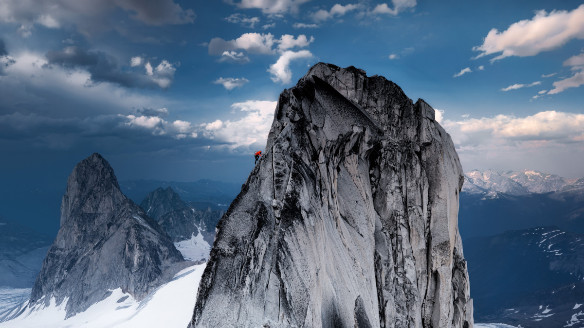A tiny climber in red stands near the summit of a jagged granite spire against a blue sky with clouds.