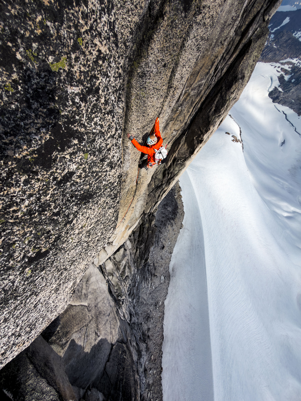A climber in an orange jacket spreads across a steep granite wall with a snow slope far below.