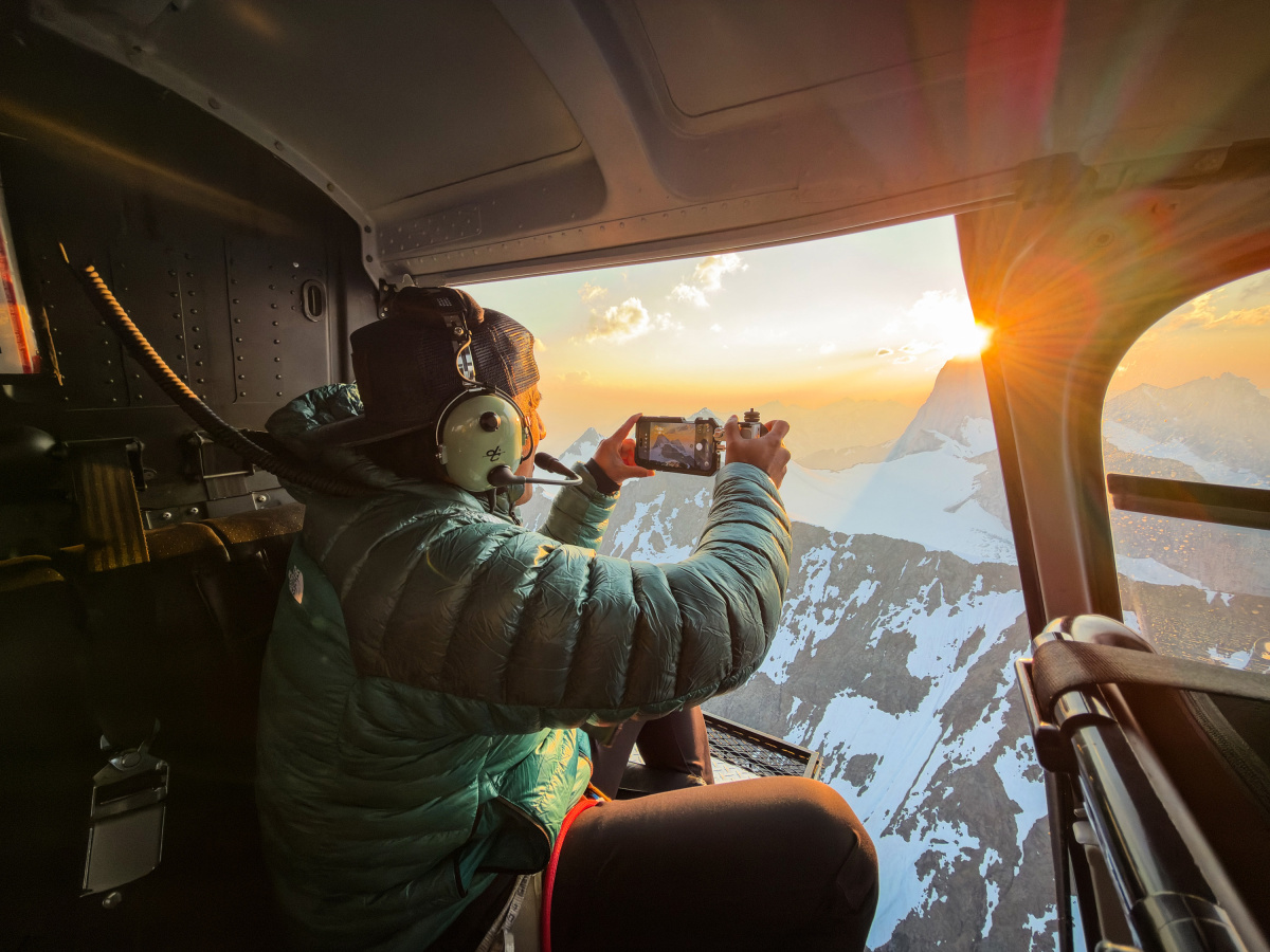 Seen from inside a helicopter, a person in a green jacket and headset holds a phone toward the open door as the sun sits low over snowy mountains.