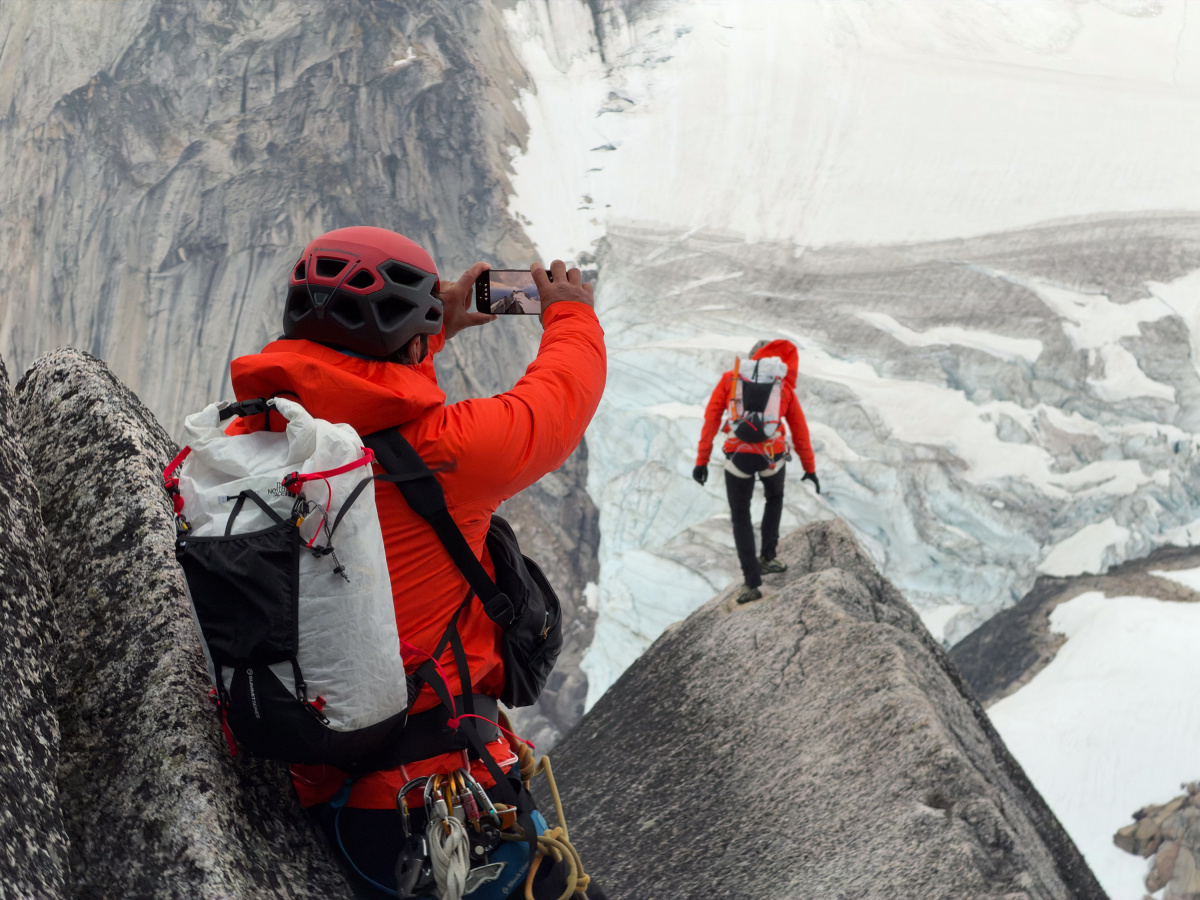 A climber in a red jacket and helmet holds up a phone to photograph a second climber walking a rocky ridge above a glacier.