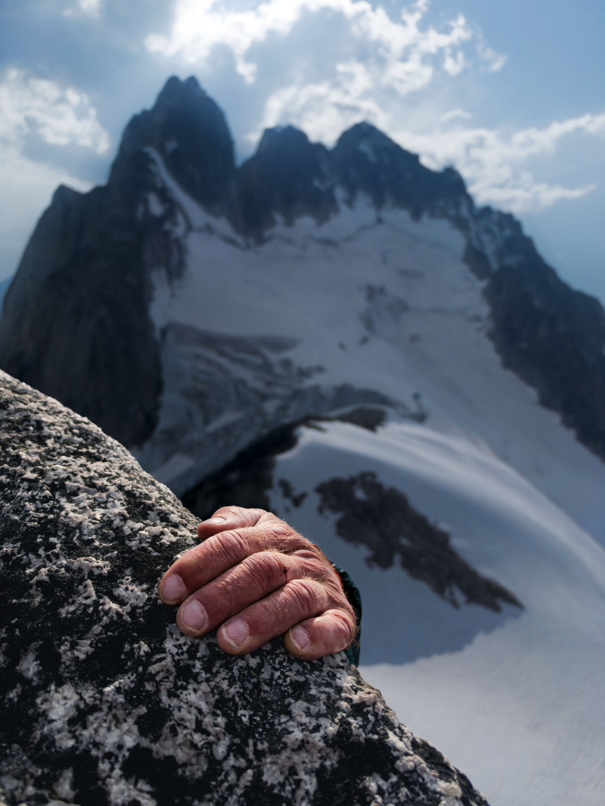 A weathered hand grips a granite edge in the foreground while snow-covered spires rise behind it.