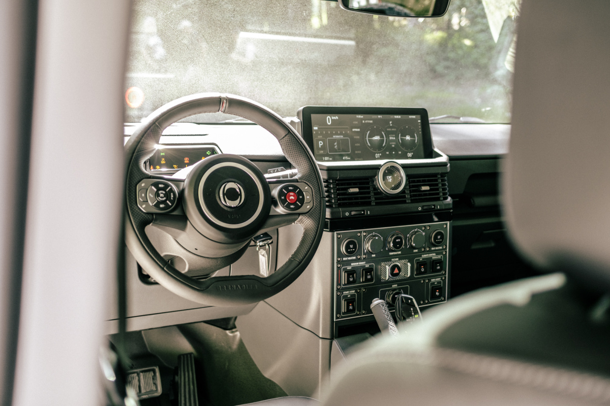 The INEOS Grenadier interior from the passenger side, showing the steering wheel with a red button on the right spoke, a rectangular touchscreen, and rows of toggle switches on the center stack.