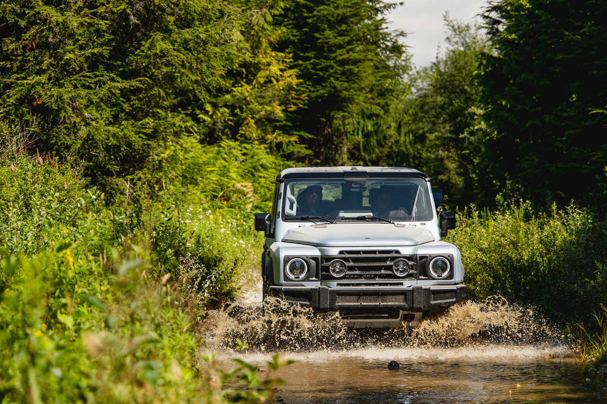 A silver INEOS Grenadier driving through a shallow stream, water spraying from the front wheels, tall green brush on both banks.