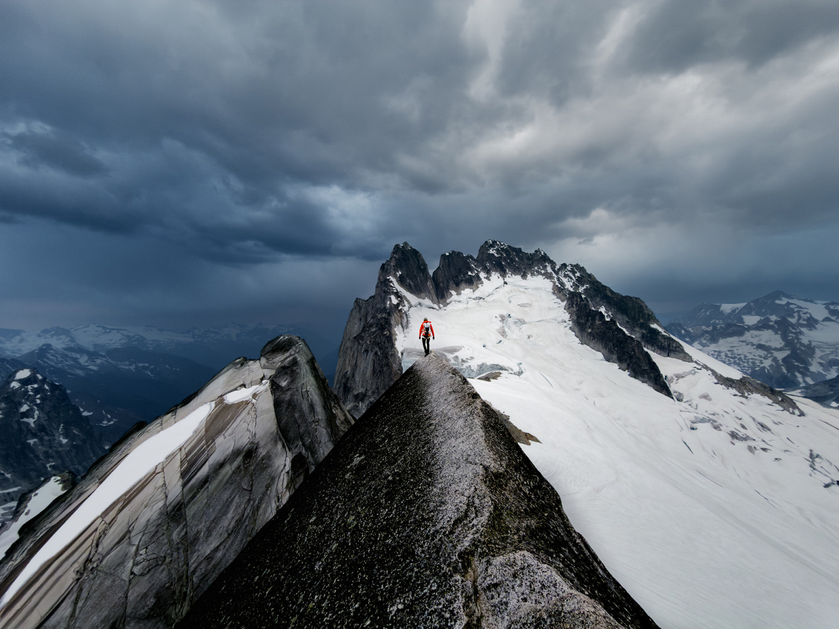 A lone climber walks a narrow rock ridge toward a snow-covered summit under dark storm clouds.