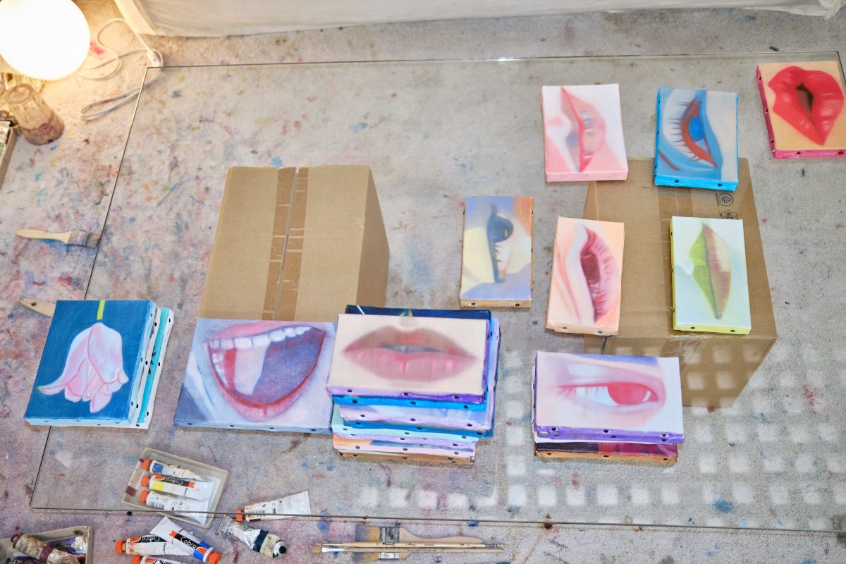 Small paintings of mouths, eyes and a flower laid out on a paint-covered studio floor beside tubes of paint.
