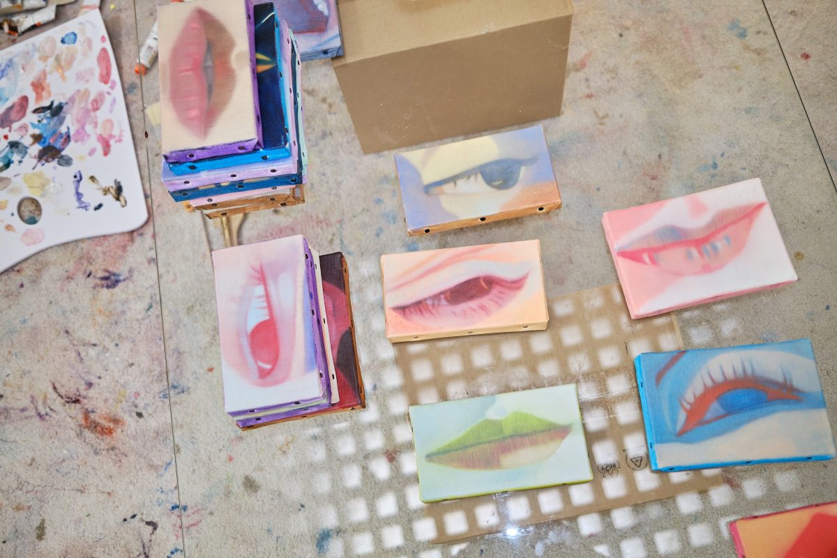 Overhead view of small paintings of lips and eyes stacked and spread across a studio floor.