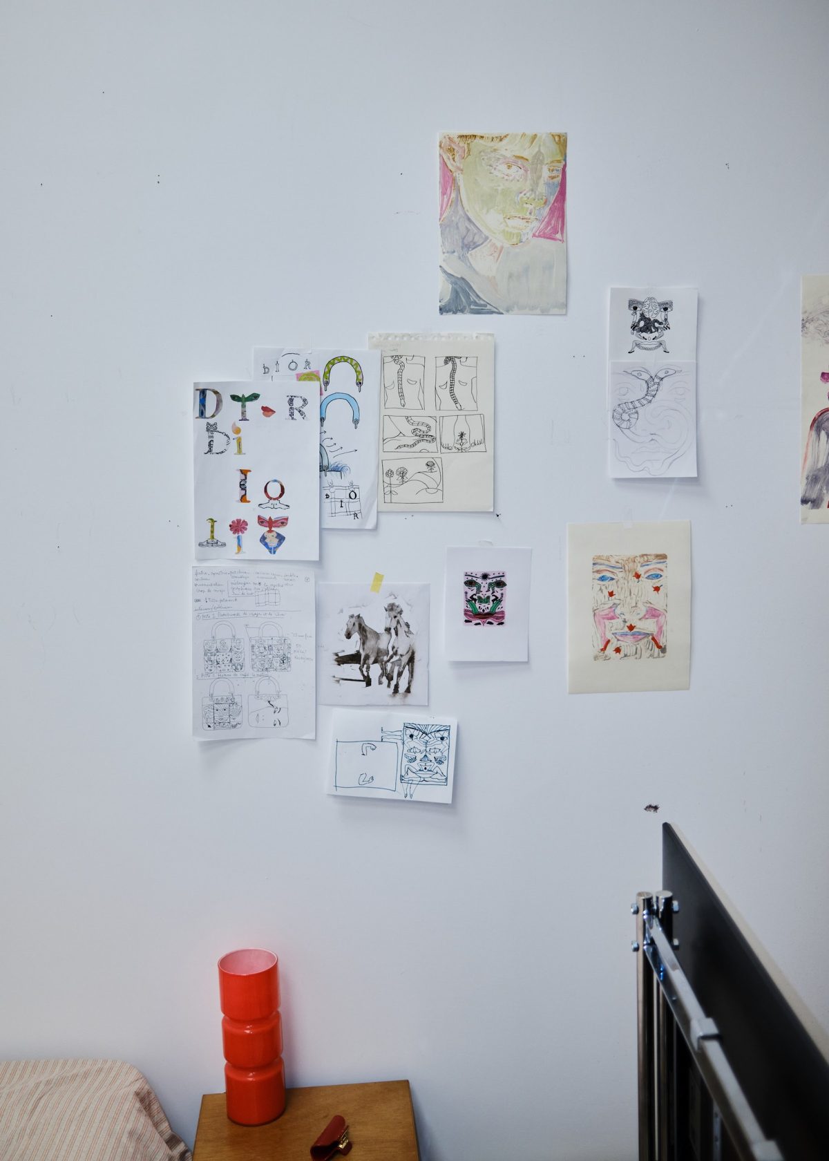 Drawings and sketches pinned to a white studio wall above a small wooden side table with a red object on it.