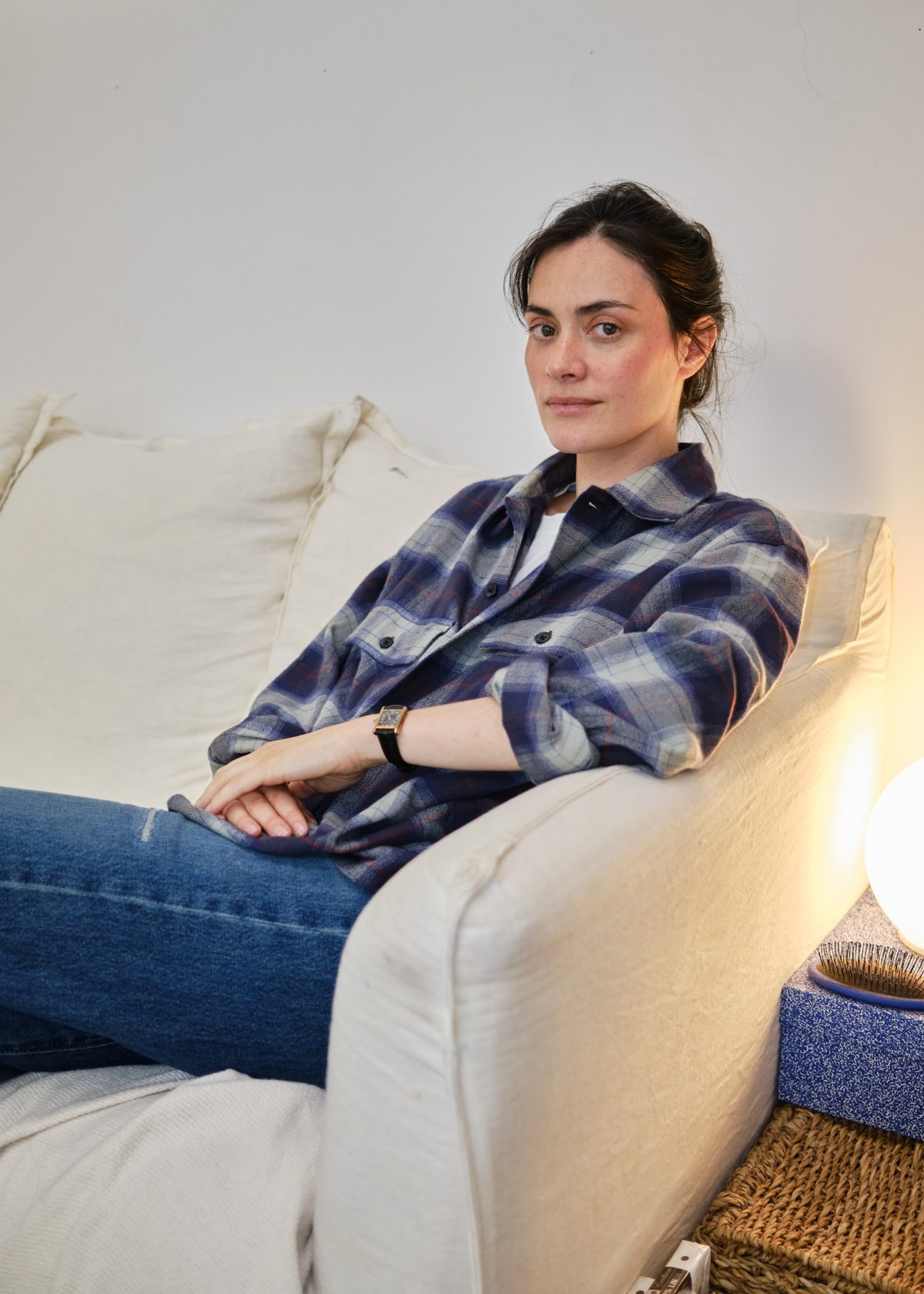 Inès Longevial in a plaid shirt and jeans sits on a white sofa with one arm along the backrest.