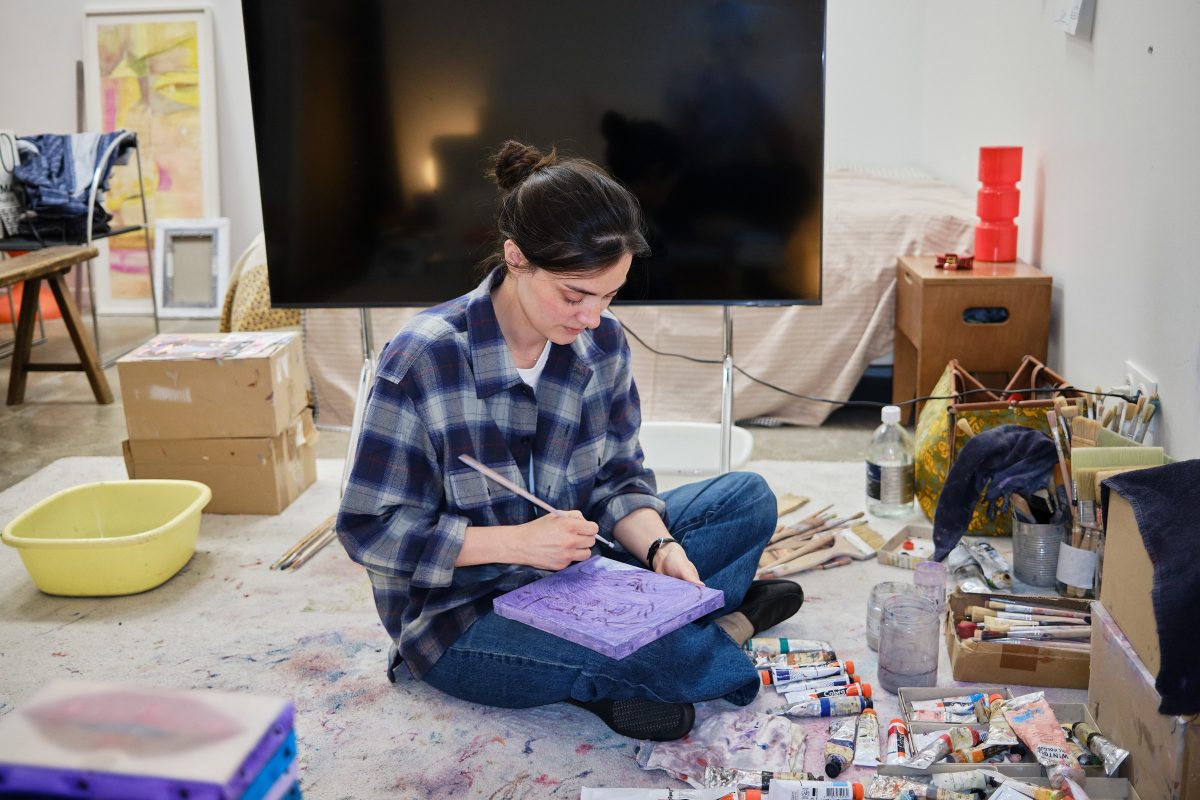 Inès Longevial sits cross-legged on a studio floor painting a small purple canvas, with boxes, a television and plastic basins around her.