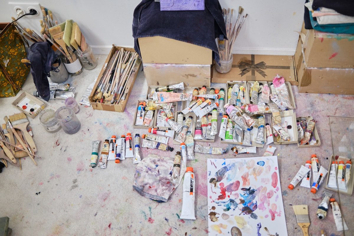 Dozens of oil paint tubes, brushes in jars and a paint-covered palette sheet spread across a studio floor.