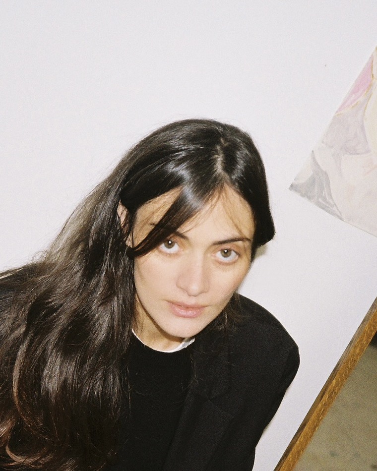 Portrait of Inès Longevial looking up at the camera, dark hair over a black jacket, in front of a white wall.