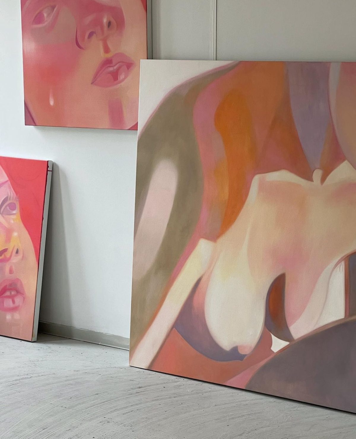 Large paintings of faces and a nude figure in pink and mauve leaning against a white studio wall.