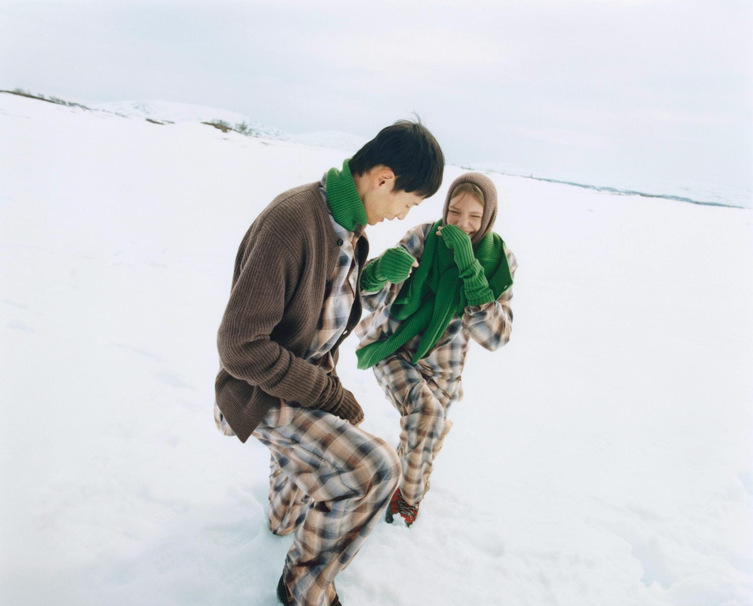 Two models in plaid flannel pajamas, a brown cardigan and green knit scarves laughing in a snowfield.