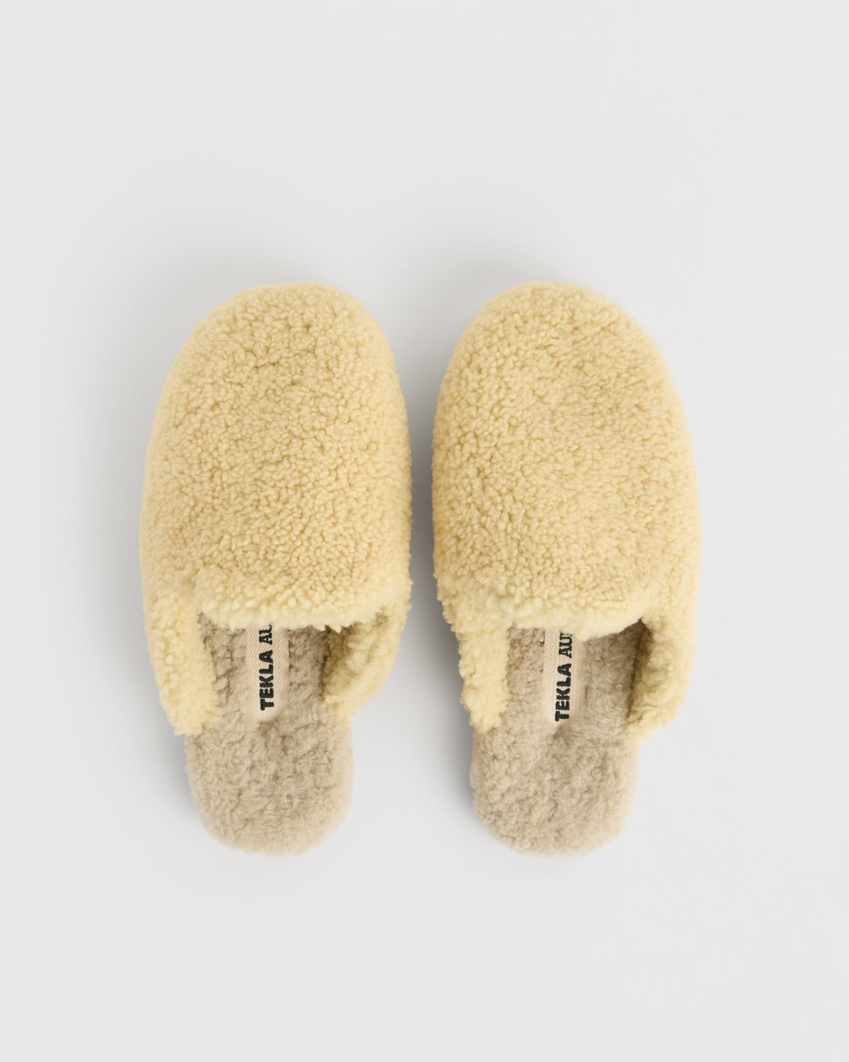 A pair of pale yellow shearling slippers seen from above on a light grey background.