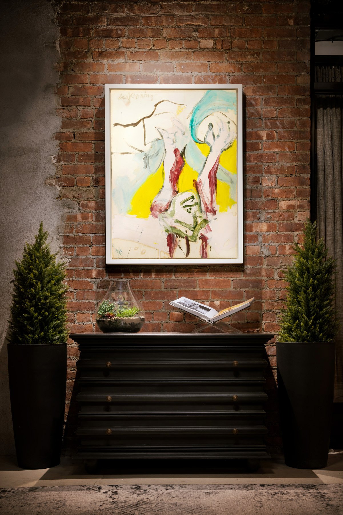 A large abstract painting in yellow, red and teal hangs on an exposed brick wall above a black chest flanked by two planters.