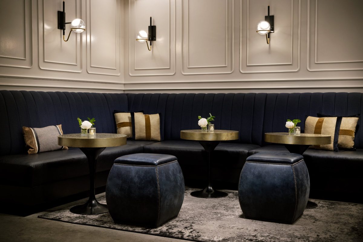 A small lounge with white paneled walls, a navy banquette, round cocktail tables and black leather drum stools.