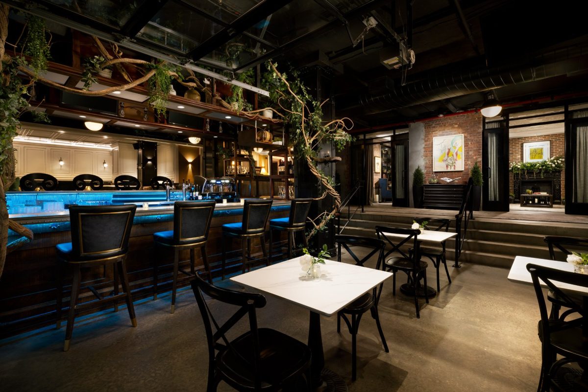 A hotel bar with a blue-lit counter, bar stools, small cafe tables and vines hanging from the ceiling.