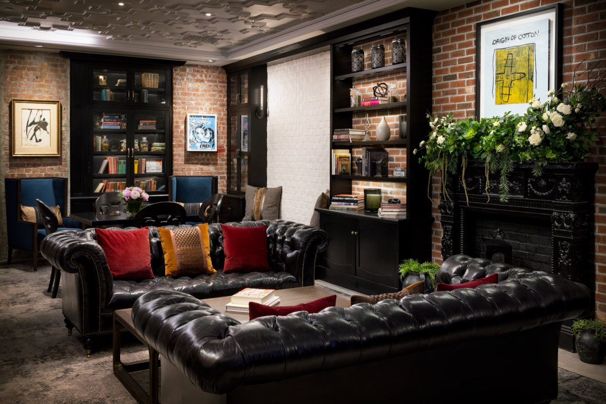 A hotel library with black leather chesterfield sofas, a fireplace, bookcases and framed artwork on brick walls.