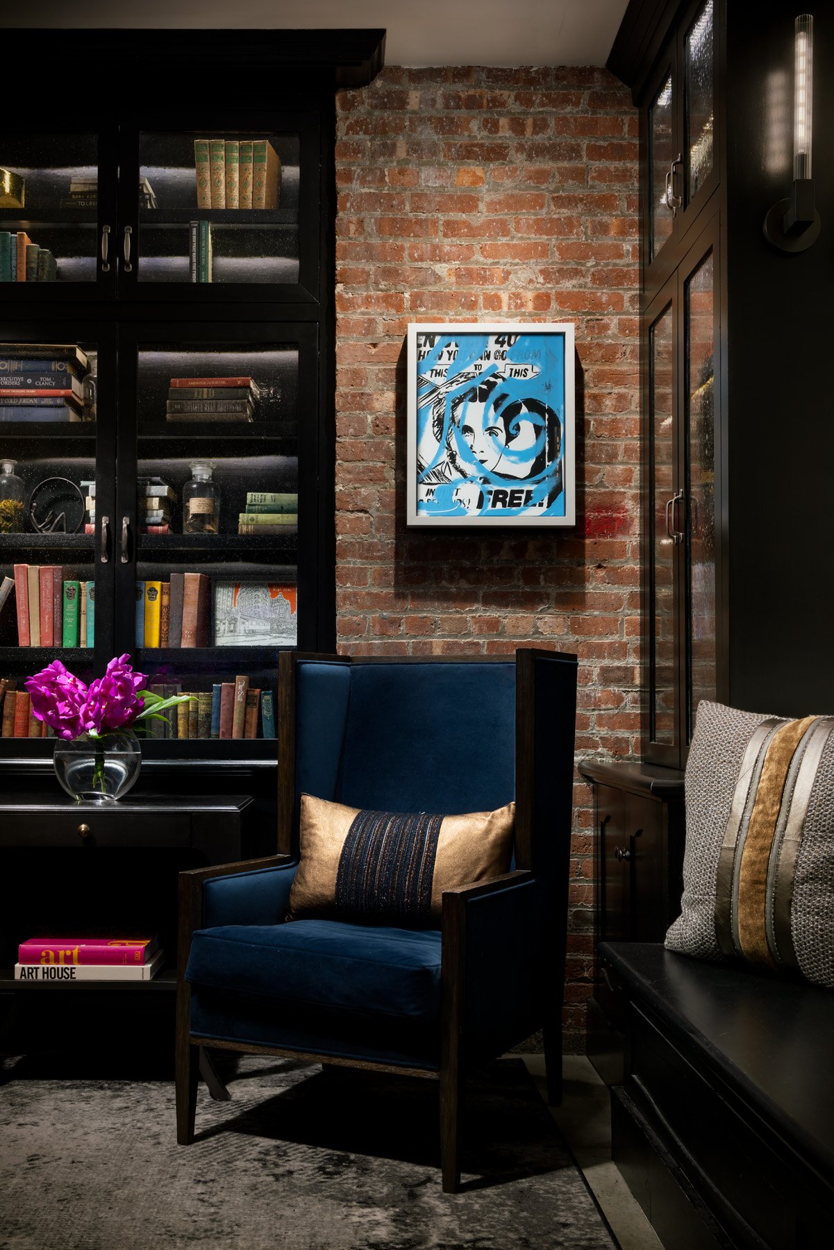 A navy velvet wingback chair beside a glass-fronted bookcase and a brick wall with a framed blue print.