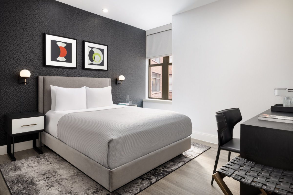 A hotel bedroom with a dark textured accent wall, a gray upholstered bed, white bedding and a black desk with a chair.