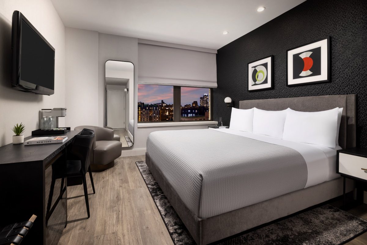 A hotel bedroom at dusk with a city view through the window, a gray bed, a desk and a wall-mounted television.