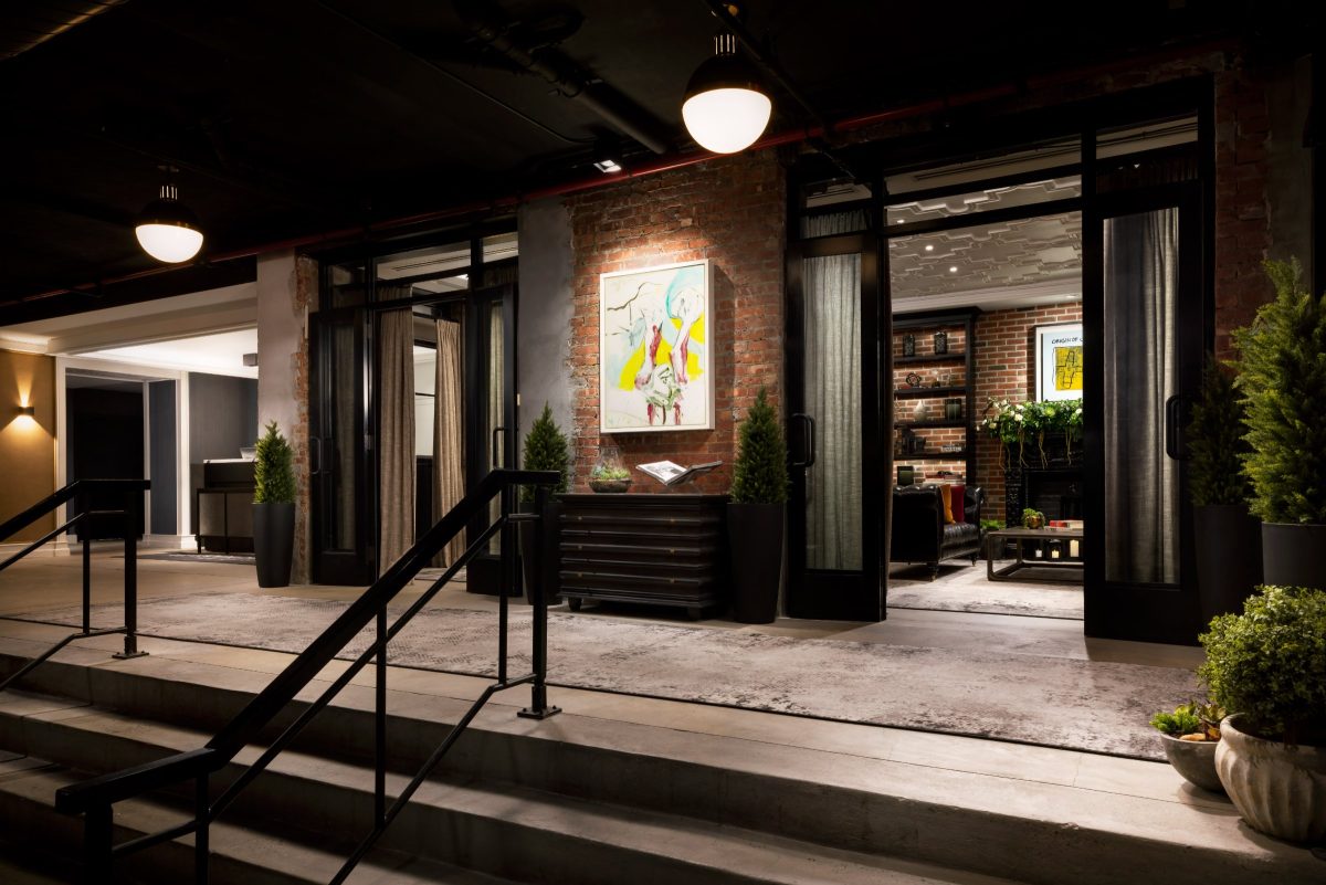 Black steel stairs rise from a hotel entrance toward a lobby with brick walls, curtains and a framed painting.