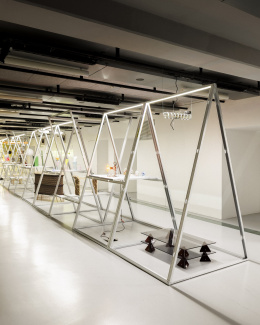 A long row of triangular steel frames lit along their peaks, with a smoked-glass table on dark pyramid feet in the foreground.