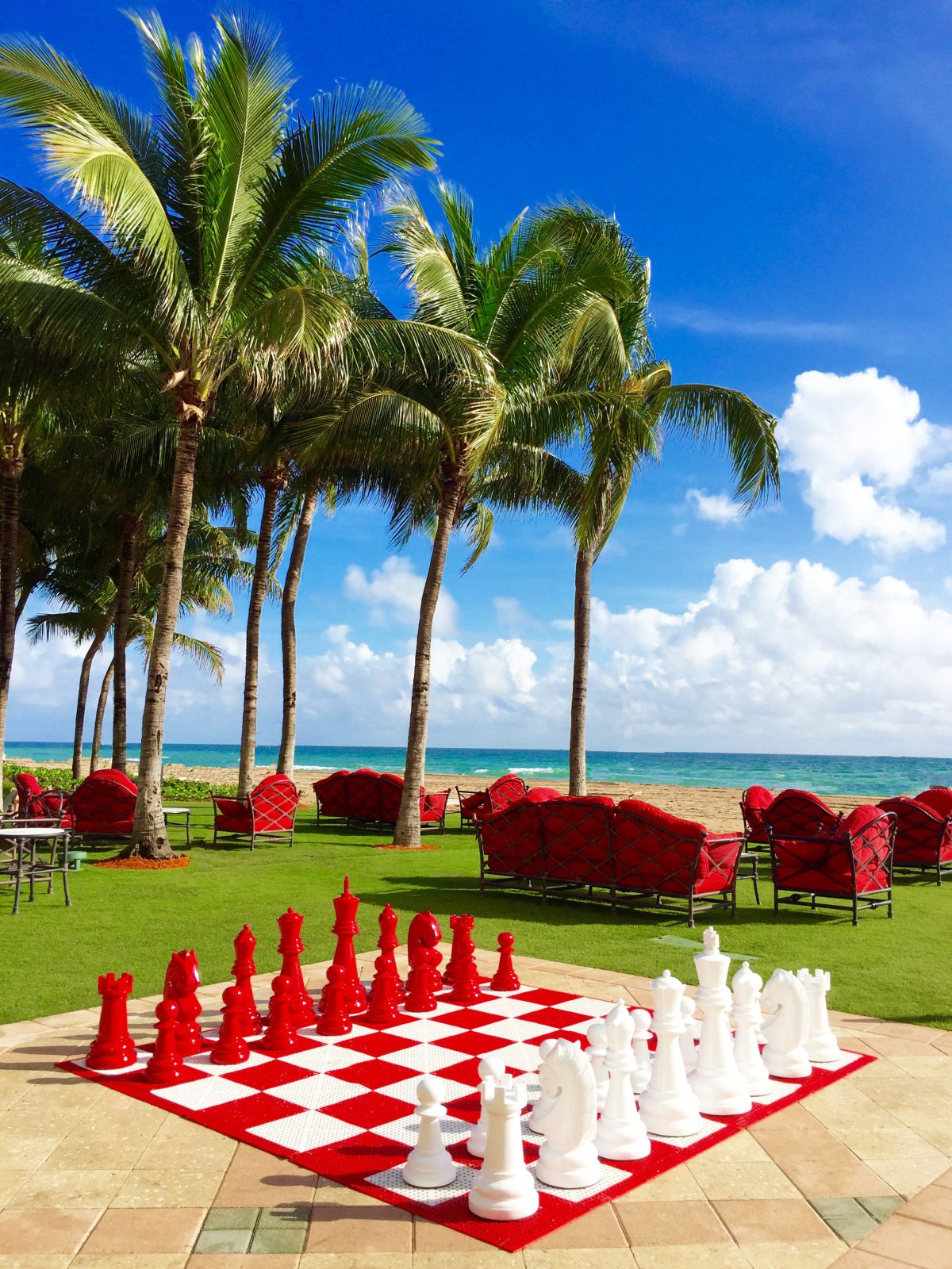 A giant red and white chess set on a lawn at Acqualina, with red seating, palm trees and the ocean beyond.