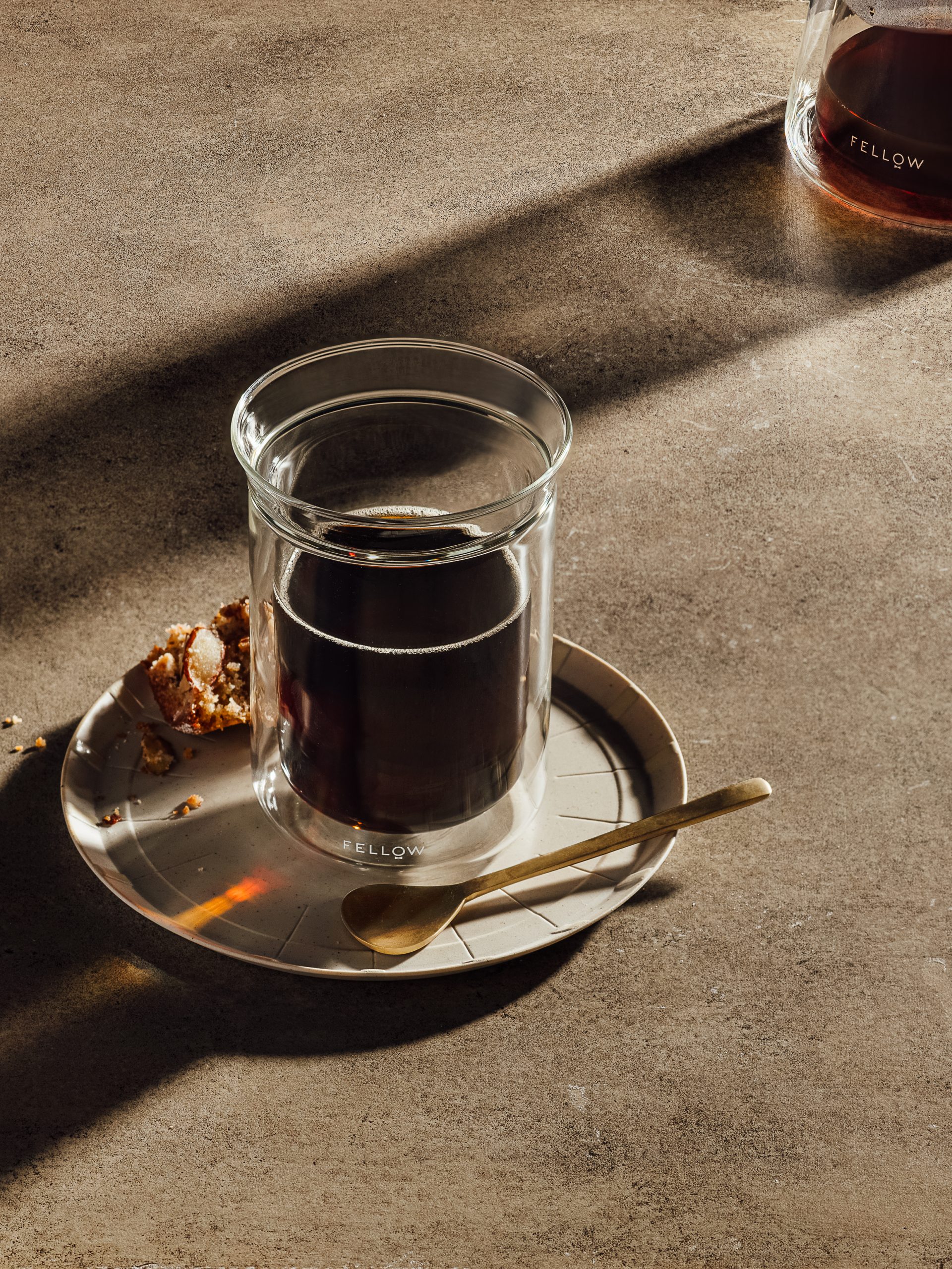 A double-walled glass of black coffee on a saucer with a gold spoon and a pastry, lit by raking sun on a stone surface.