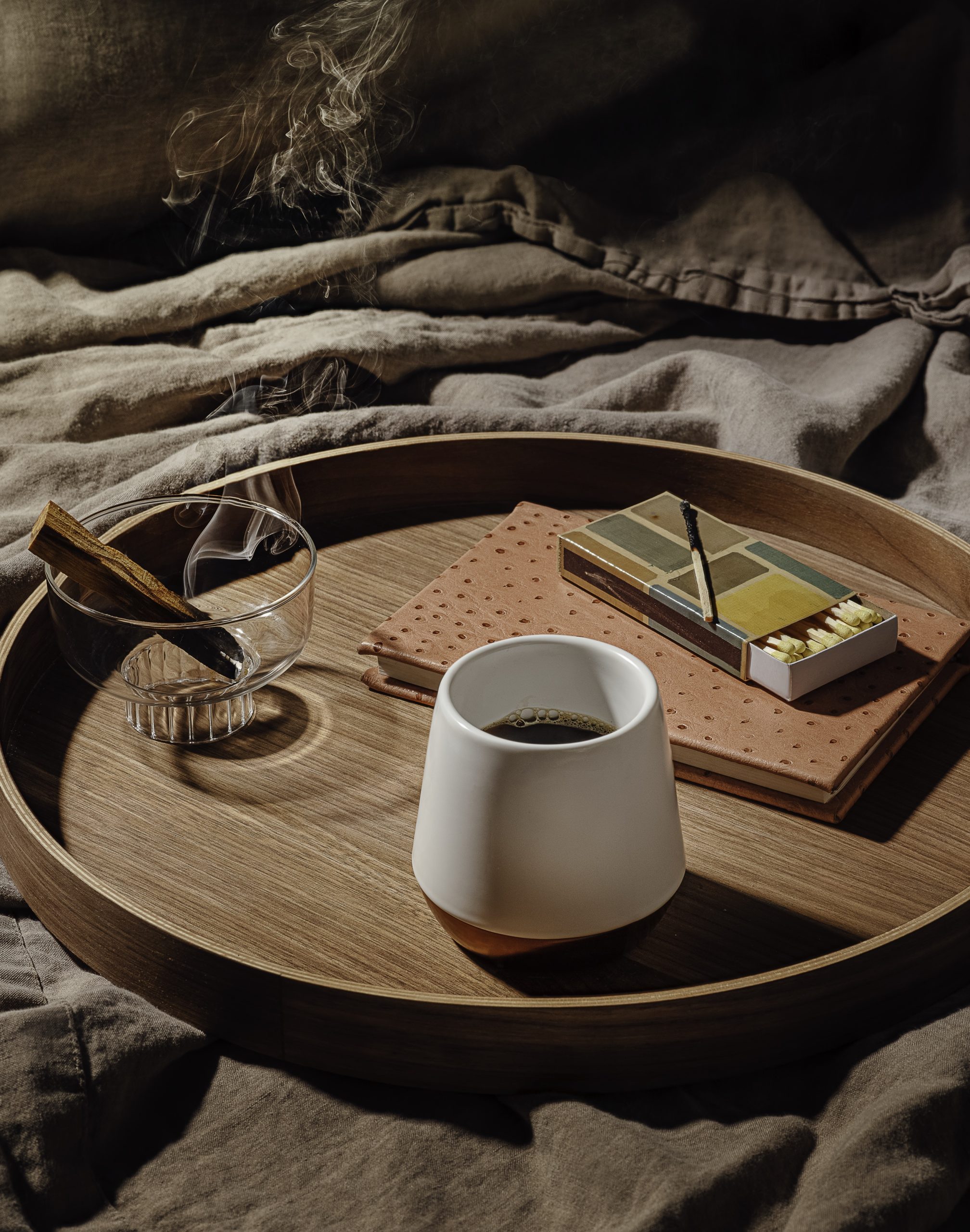 A white cup of coffee on a round wood tray with a glass and a box of matches, set on rumpled gray linen.
