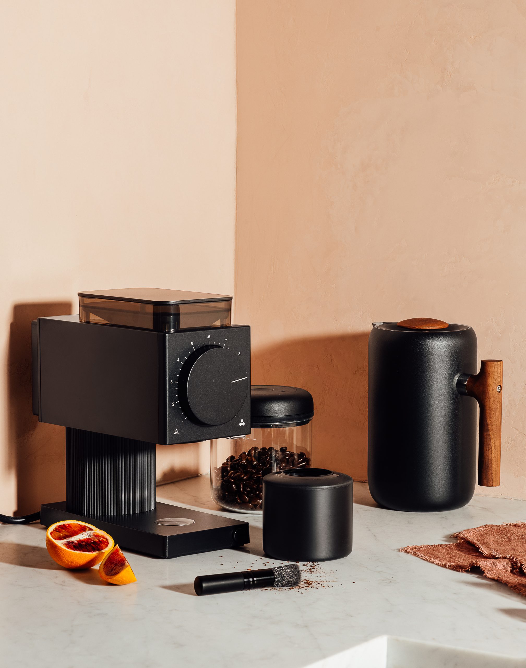 A black Fellow coffee grinder, a jar of beans and a black pitcher with a wood handle on a marble counter beside a halved orange.