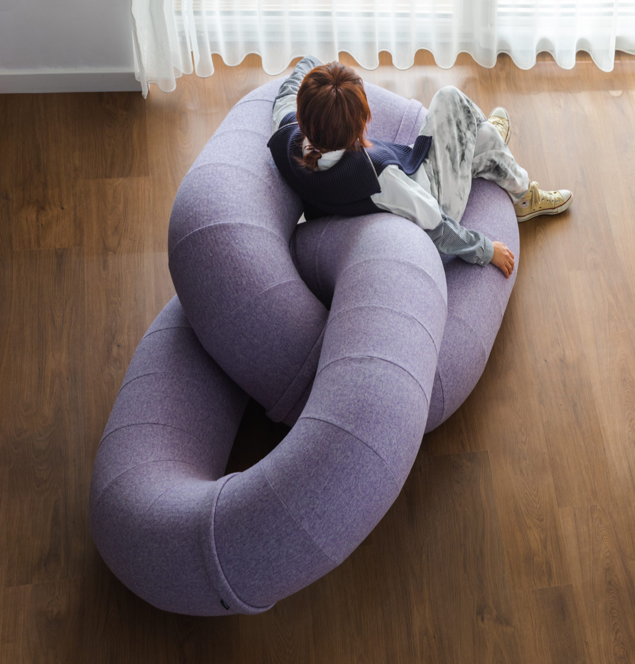 A person lounges on a lavender sofa made of linked upholstered loops on a wood floor.
